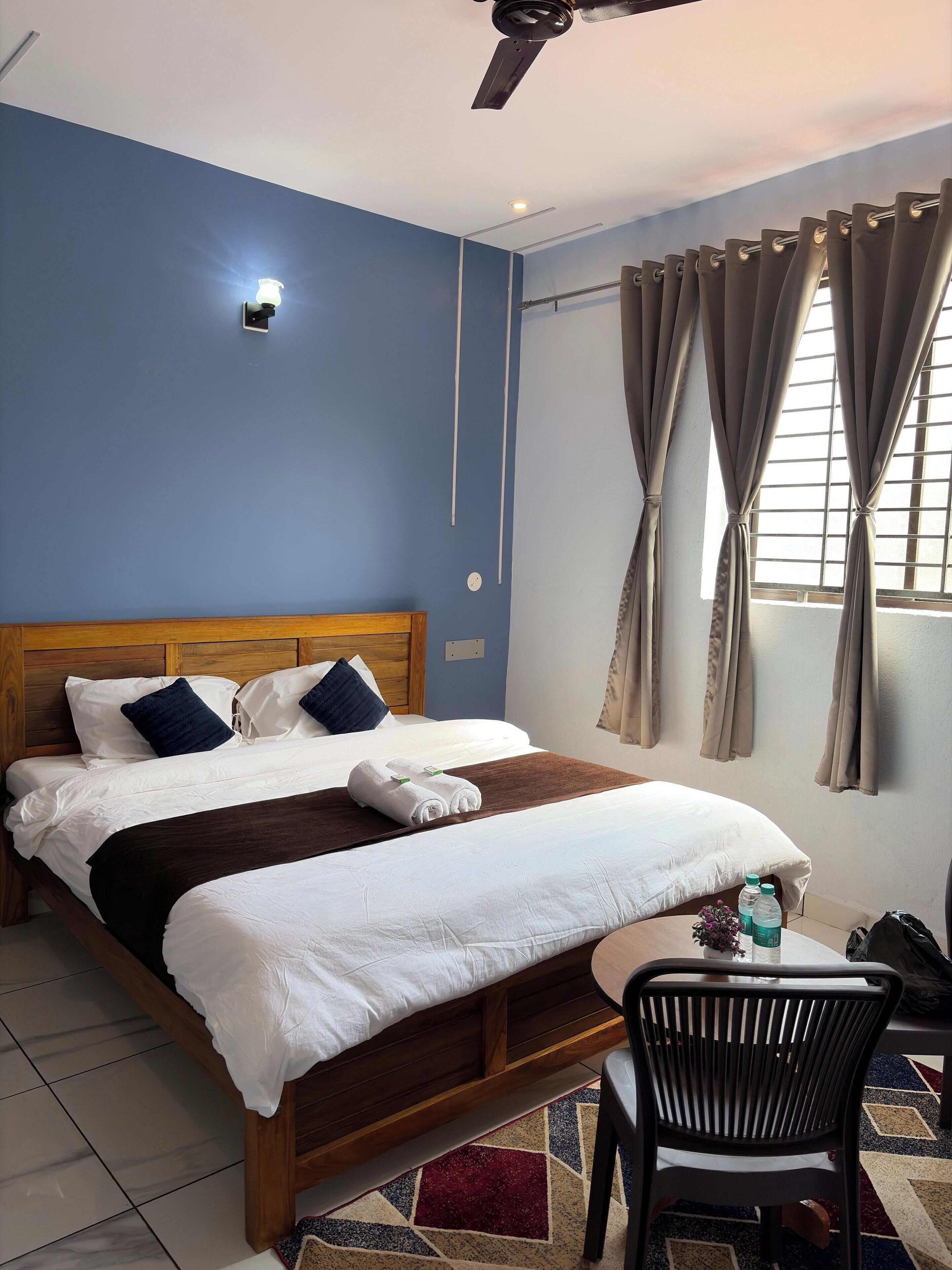 Camera d'albergo con un letto di legno, una ventola a soffitto, una finestra con tende e un piccolo tavolo con una sedia.