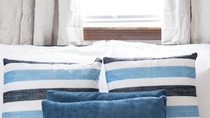 A bed with white bedding and two pillows, one striped and one solid blue, in front of a window with white curtains. - Near K-State Campus - Central Manhattan Stay (Manhattan)