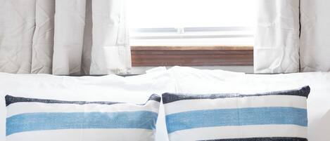 Una cama con ropa de cama blanca y dos almohadas, una a rayas y otra azul lisa, frente a una ventana con cortinas blancas.