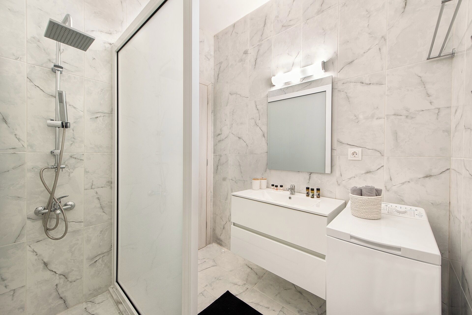 Une salle de bain moderne dotée d’une douche à parois de verre, d’un grand miroir et d’un meuble-lavabo blanc avec un évier et un plan de travail.