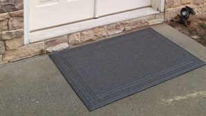 Une porte à deux battants, un mur en pierre à la texture particulière, un paillasson et un petit espace extérieur agrémenté d’un objet en métal.