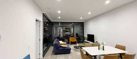 A modern living room with a sofa, dining table, and chairs. There is a TV and a painting on the wall.