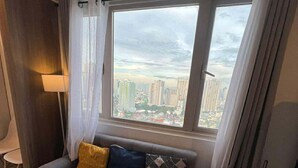 A modern living room with a grey sofa, two cushions, a small round table, and a view of a cityscape through large windows. - Bayfront Cozy Stay at SMDC Coast Residences (Pasay City)