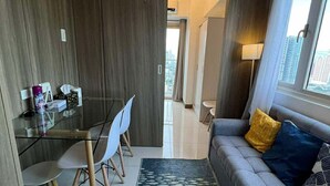 A modern living room with a grey sofa, a dining table with chairs, and a city view through large windows. - Bayfront Cozy Stay at SMDC Coast Residences (Pasay City)