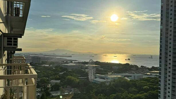 A high-rise building with a view of the sunset over a body of water and surrounding greenery. - Bayfront Cozy Stay at SMDC Coast Residences (Pasay City)