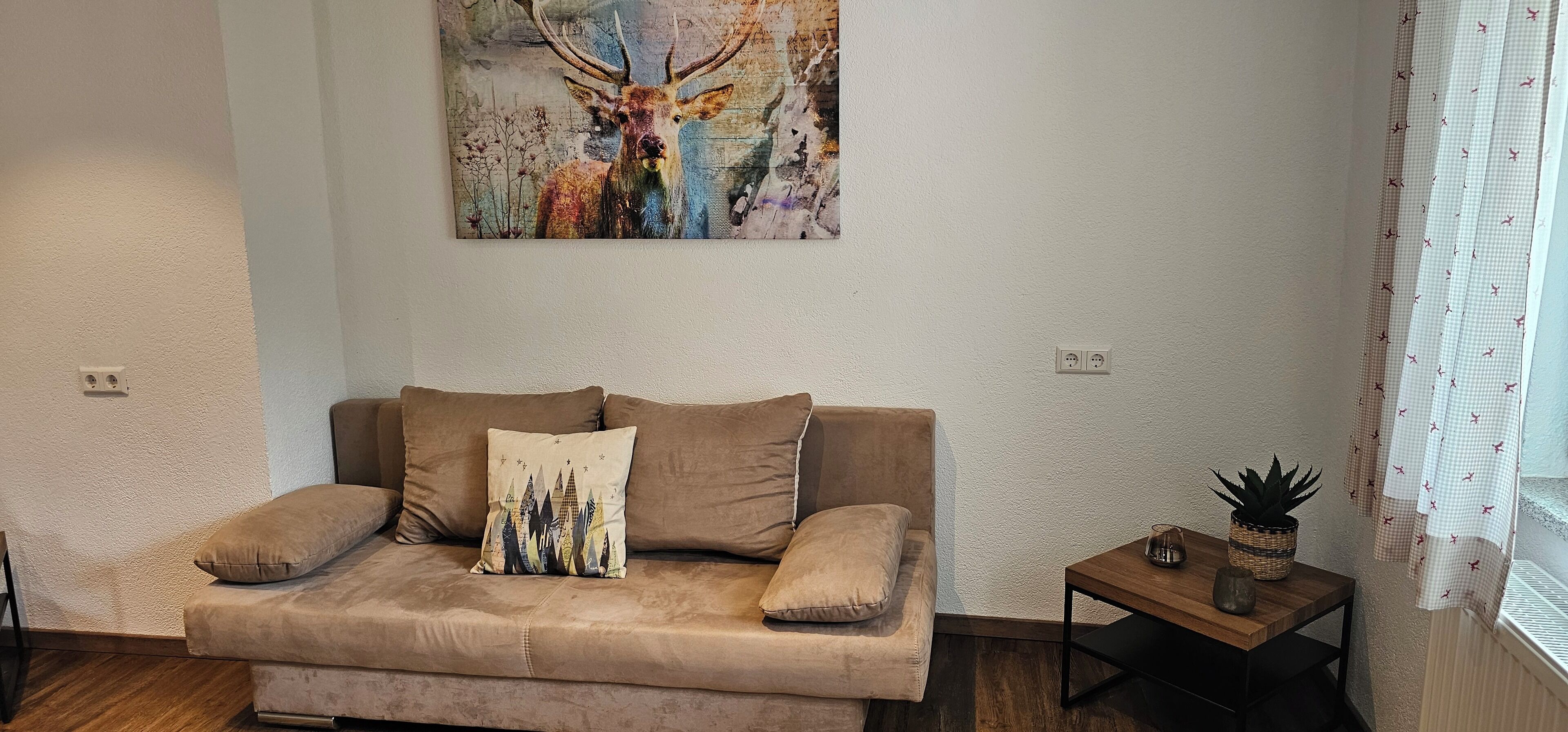 A living room with a beige sofa, a cityscape pillow, a small table with a plant, and a painting of a deer on the wall.