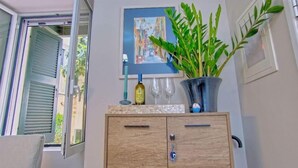 A wooden cabinet with a wine bottle and glasses on top, a potted plant, and a painting on the wall. - Indigo Apartment by Corfuescapes  (Corfu)