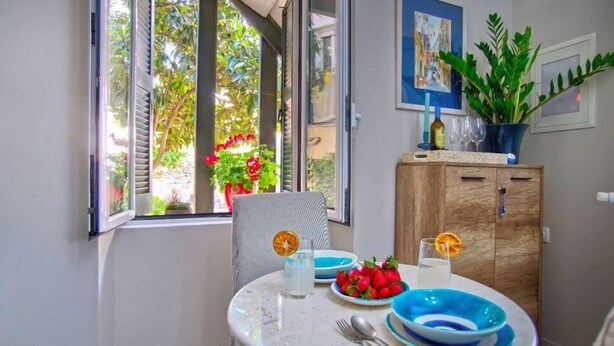 A dining area with a table set for two, featuring blue and white plates, a glass of water, and a glass of white wine. There is a wooden cabinet with a plant on top, and a window with a view of greenery outside. - Indigo Apartment by Corfuescapes  (Corfu)