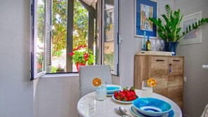 A dining area with a table set for two, featuring blue and white plates, a glass of water, and a glass of white wine. There is a wooden cabinet with a plant on top, and a window with a view of greenery outside. - Indigo Apartment by Corfuescapes  (Corfu)