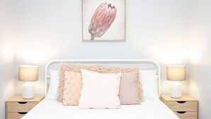 Una cama bien hecha con ropa de cama blanca, flanqueada por dos mesitas de noche con lámparas, y un cuadro de una flor colgado en la pared.