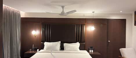 A hotel room with a large bed, a ceiling fan, two bedside tables with lamps, a wooden wardrobe, and a wooden chair.
