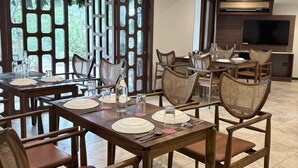 A dining area with wooden tables and chairs, a large window with a view of greenery, and a flat-screen TV mounted on the wall. -  Lords Riverside Resort Muduba - A Wellness Nature Retreat (Tirthalli)