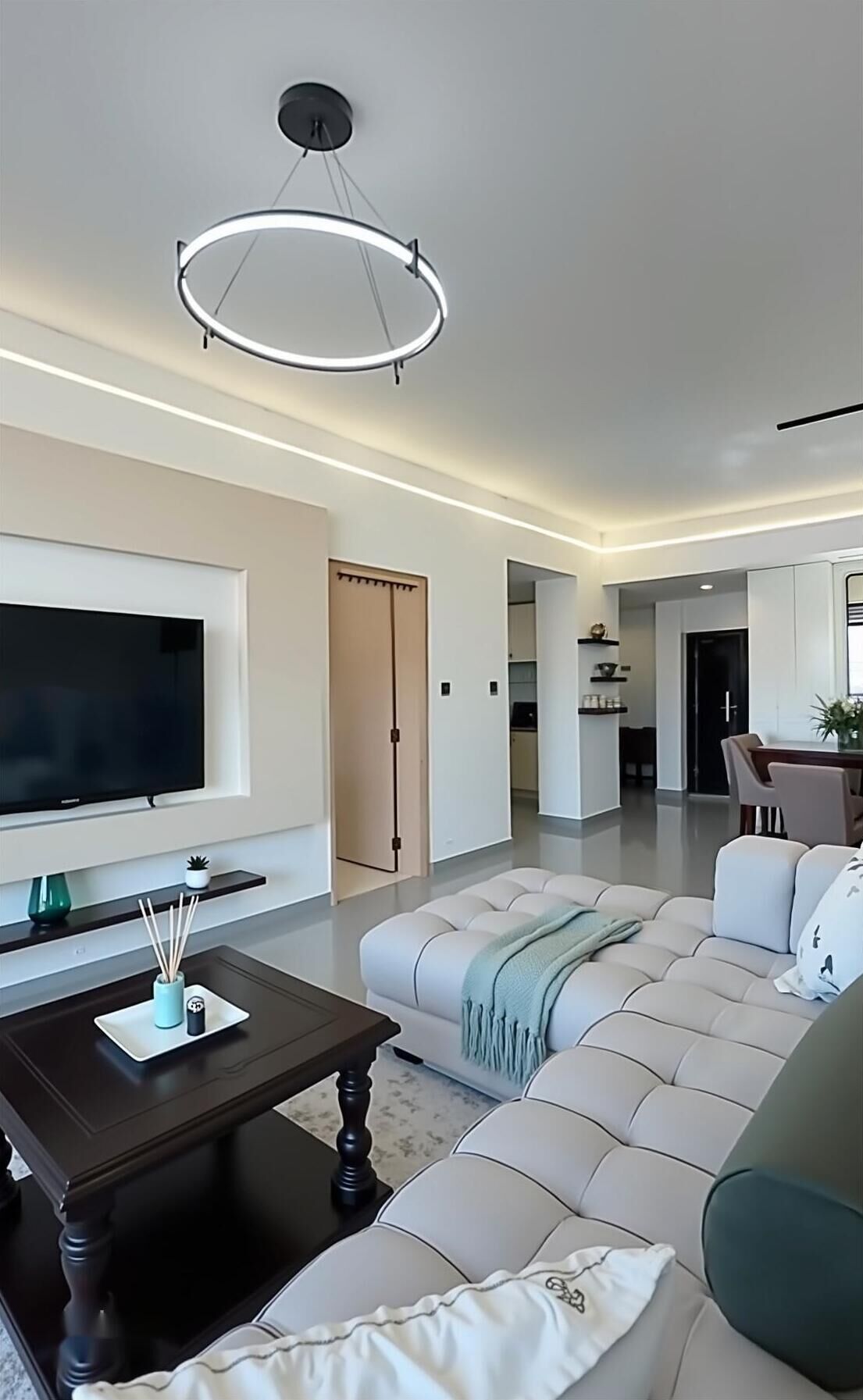 A modern living room with a large flat-screen TV, a dark wooden coffee table, a beige sectional sofa, and a dining area in the background.
