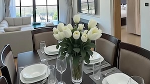 Un'area pranzo moderna con un tavolo apparecchiato per quattro persone, un vaso di fiori bianchi e vista sul soggiorno e sul balcone.