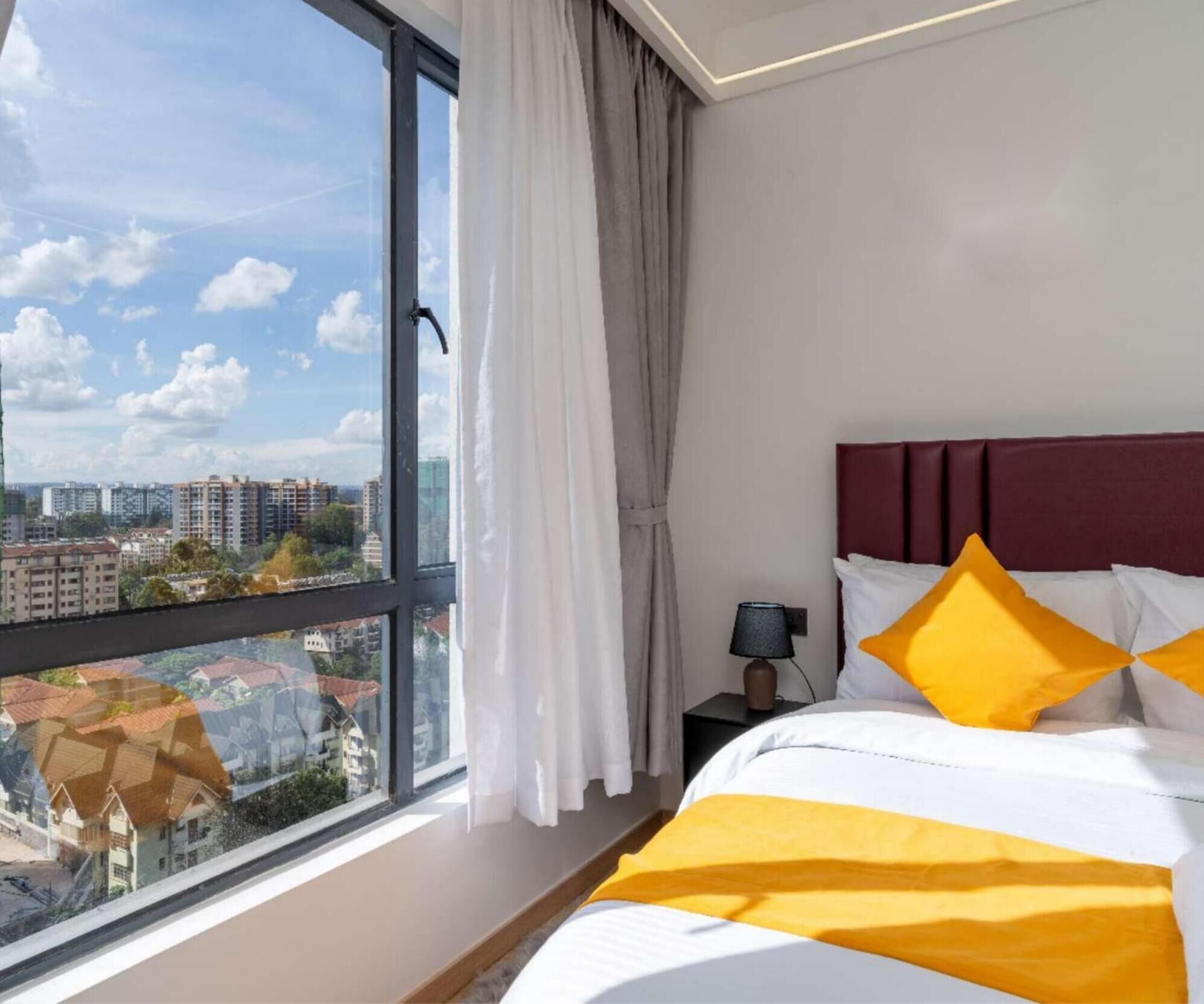 A hotel room with a large window, a bed with white and yellow bedding, a dark headboard, and a nightstand with a lamp.