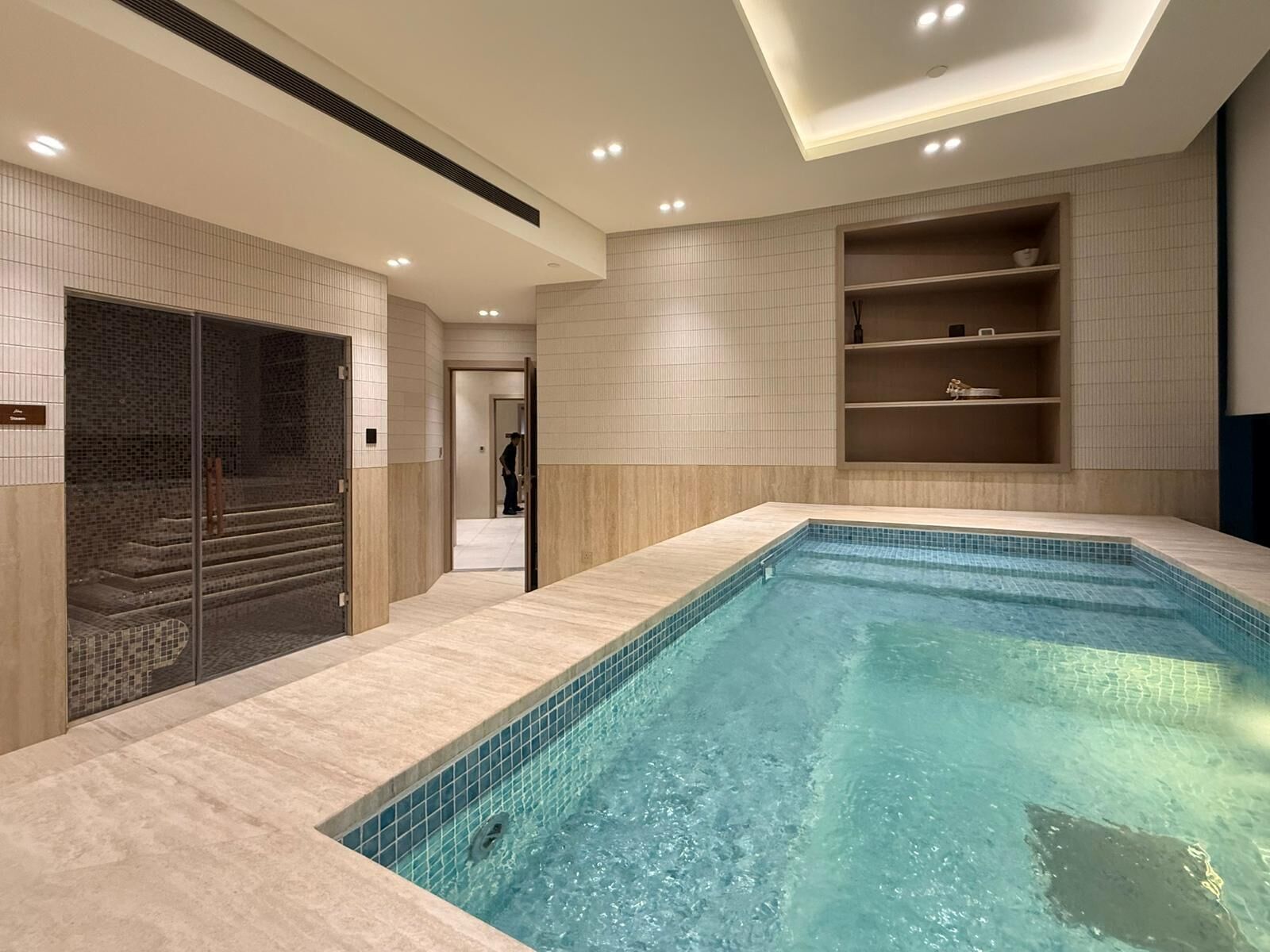 A modern indoor swimming pool with a tiled floor and walls, a glass door entrance, and built-in shelves with toiletries.