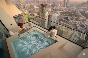 Una pareja sentada junto a una piscina infinita con cerramiento de vidrio y vista a la ciudad, con jacuzzi y un área de comedor con velas y flores.