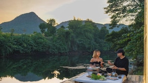 A wooden deck by a calm river with a mountain in the background. - Nubdao Glamping (Kanchanaburi)