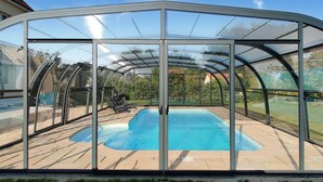 Una piscina cubierta con cerramiento de vidrio, rodeada por una terraza de madera y un césped.