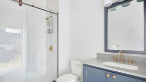 A modern bathroom with a glass shower enclosure, a white toilet, a sink with a mirror, and blue cabinets. - Two Stylish Suites + Full Kitchen | Canal St + Quarter | The Harlequin (New Orleans)