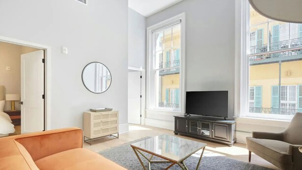 A modern living room with a sofa, a glass coffee table, a TV on a cabinet, and a view of a building with balconies. - Two Stylish Suites + Full Kitchen | Canal St + Quarter | The Harlequin (New Orleans)