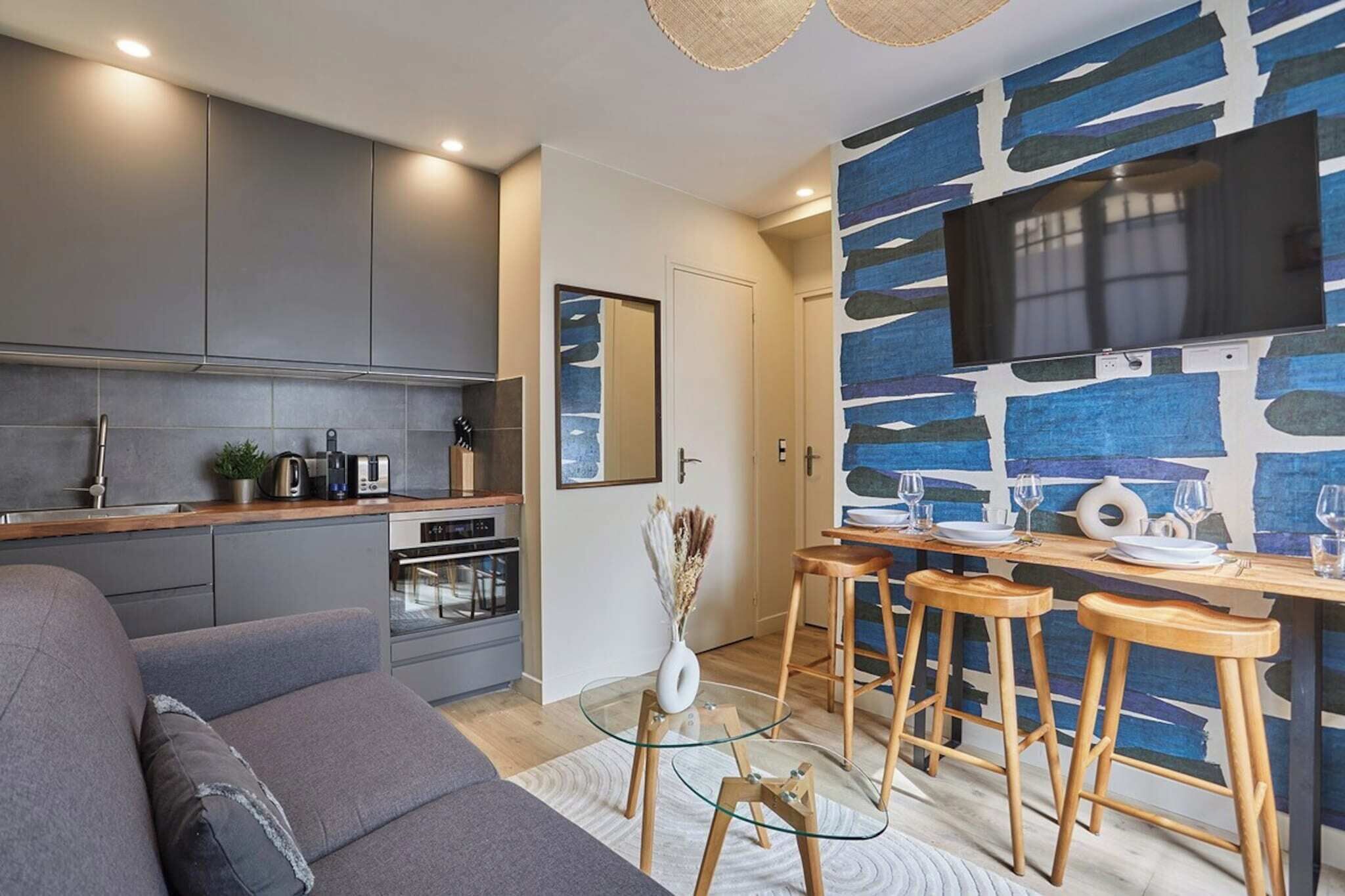 A modern kitchen with grey cabinets, a built-in oven, and a dining area with stools and a glass table.