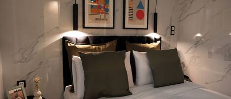 A modern bedroom with a marble wall, a bed with white bedding and multiple pillows, a nightstand with a vase and picture frame, and two framed artworks above the bed.