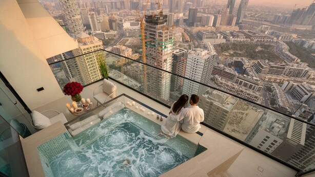 Una coppia seduta accanto a una piscina a sfioro con pareti in vetro e vista sulla città, con idromassaggio e zona pranzo con candele e fiori.