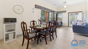 A dining area with a wooden table and chairs, a microwave on a shelf, and a large window with a view of greenery.