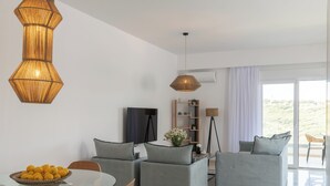 En moderne stue med et glasbord, grå sofaer, en fletvæglampe og et stort vindue med udsigt til grønne områder.