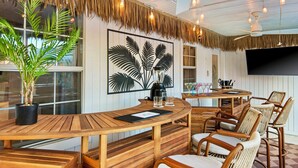 A tropical-themed room with a wooden dining table, wicker chairs, a large potted plant, and a wall-mounted TV. - PCB Stay w/ Heated Pool, Hot Tub, Bars, Game Room & Quick Walk to Beach (Panama City Beach)