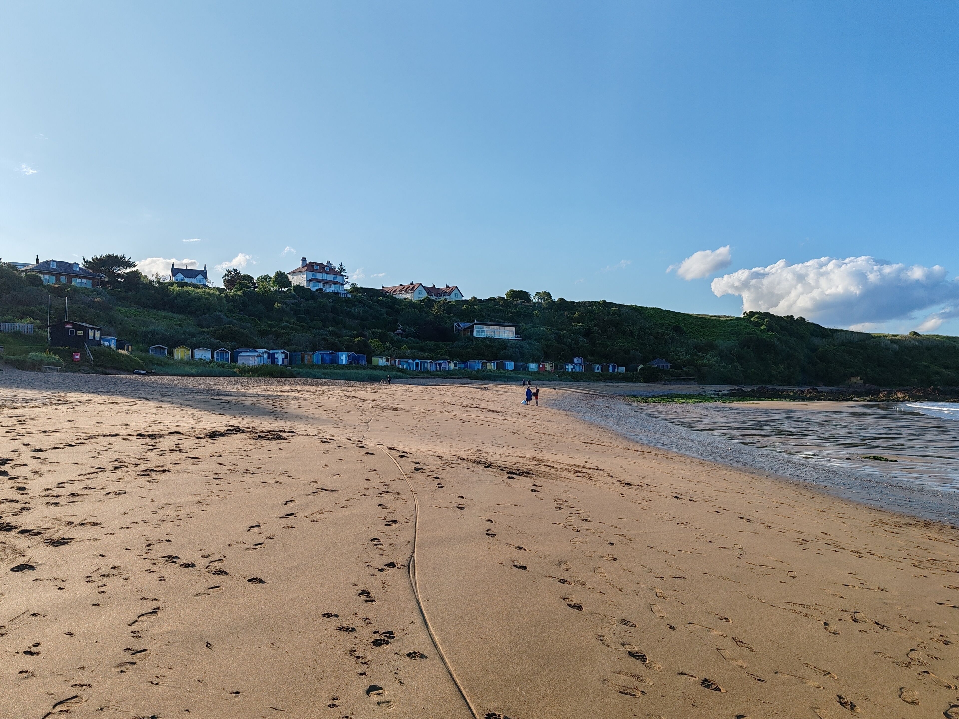 Une plage de sable où l’on peut voir des empreintes menant à l’eau, des maisons sur une colline et un ciel dégagé.
