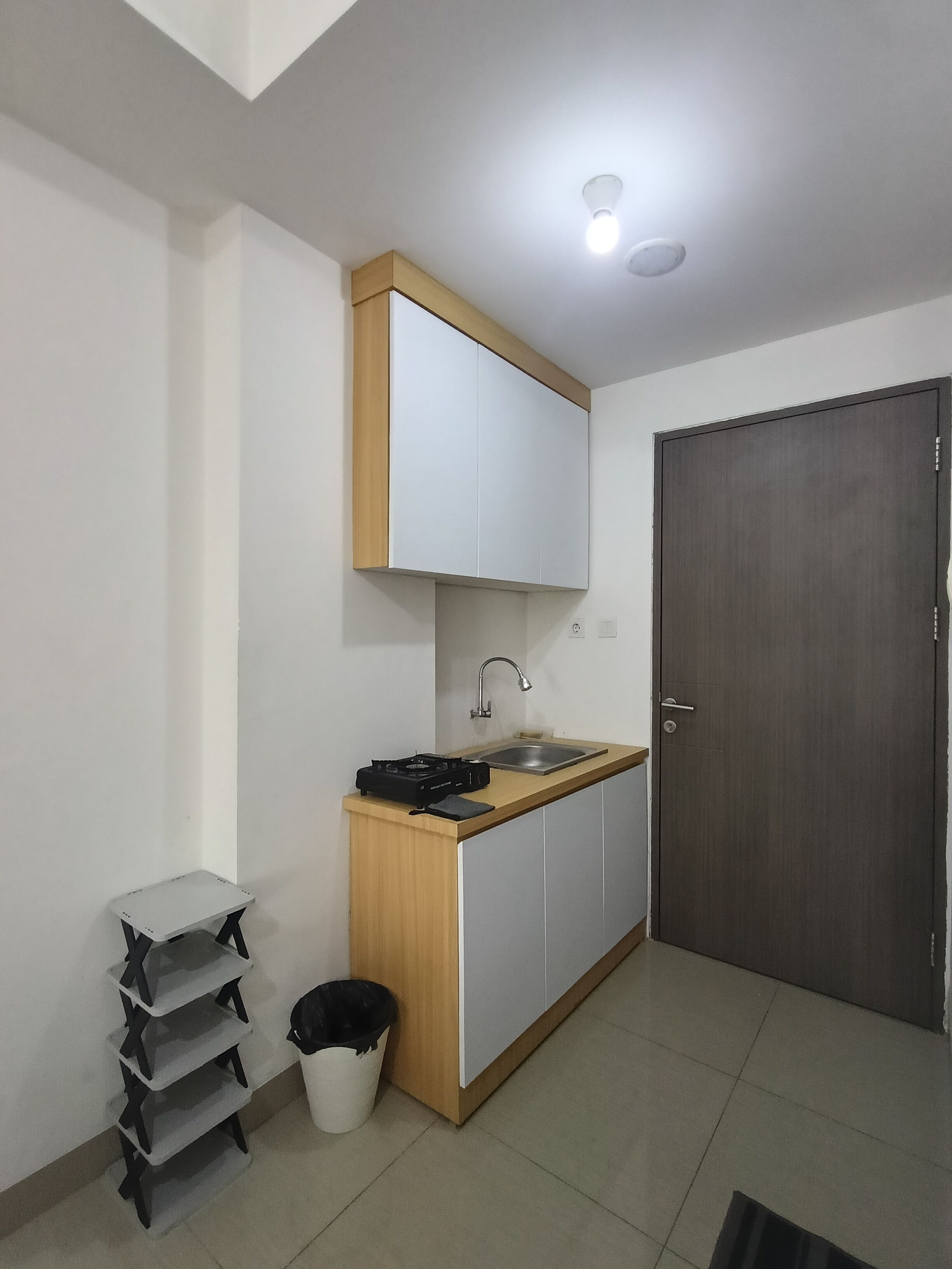 Een kleine kitchenette met een spoelbak, een kookplaat en een kastje boven het aanrecht. Er is een prullenbak en een klein tafeltje met planken.