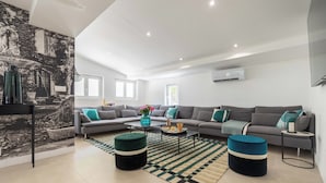 Un salon moderne avec un canapé d’angle, une table basse et deux poufs. Une grande fenêtre laisse entrer la lumière naturelle et un tableau mural décore le mur.
