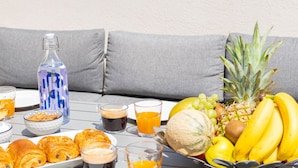 Une table dressée avec des aliments pour le petit-déjeuner, notamment des croissants, des fruits et des boissons, sur une terrasse extérieure d’où l’on peut admirer une ville et des montagnes en arrière-plan.