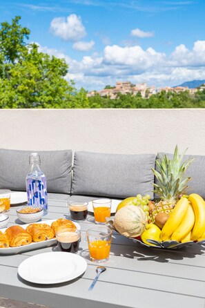 A table set with breakfast items, including croissants, fruit, and beverages, on an outdoor patio with a view of a town and mountains in the background. - Mas de l'Etoile - Exceptional villa in Provence (Mimet)