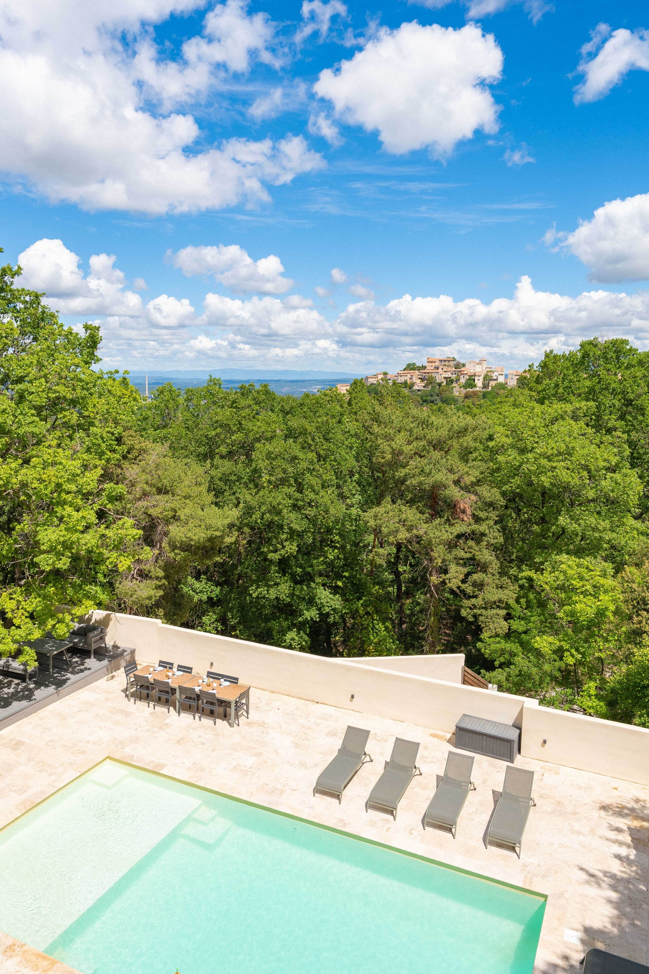 A rooftop pool with lounge chairs, a dining area, and a view of a forested area with a distant building.