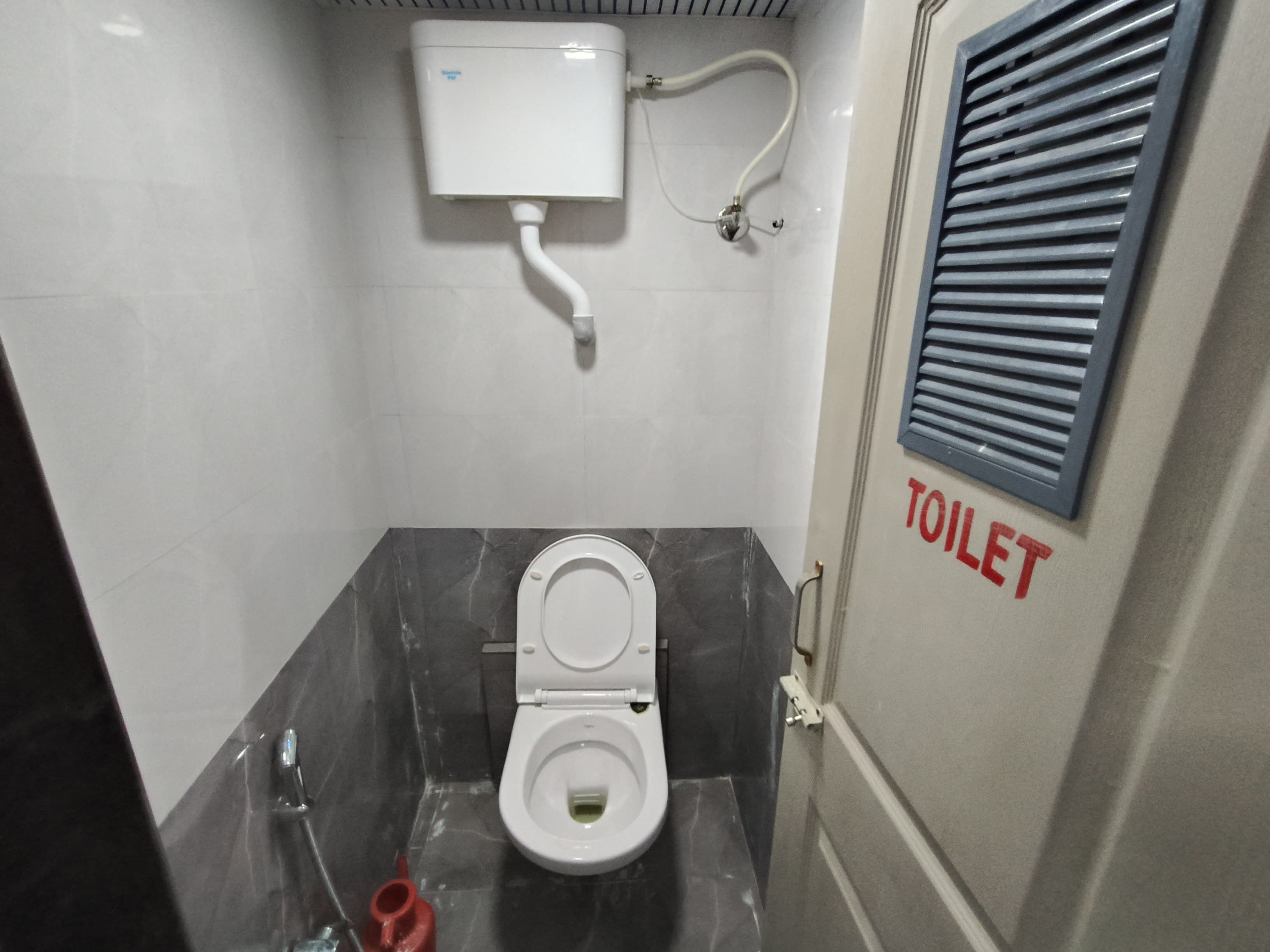 Une petite salle de bain moderne, équipée de toilettes, dont la tuyauterie est visible, et sur laquelle est apposée une signalétique indiquant son emplacement.