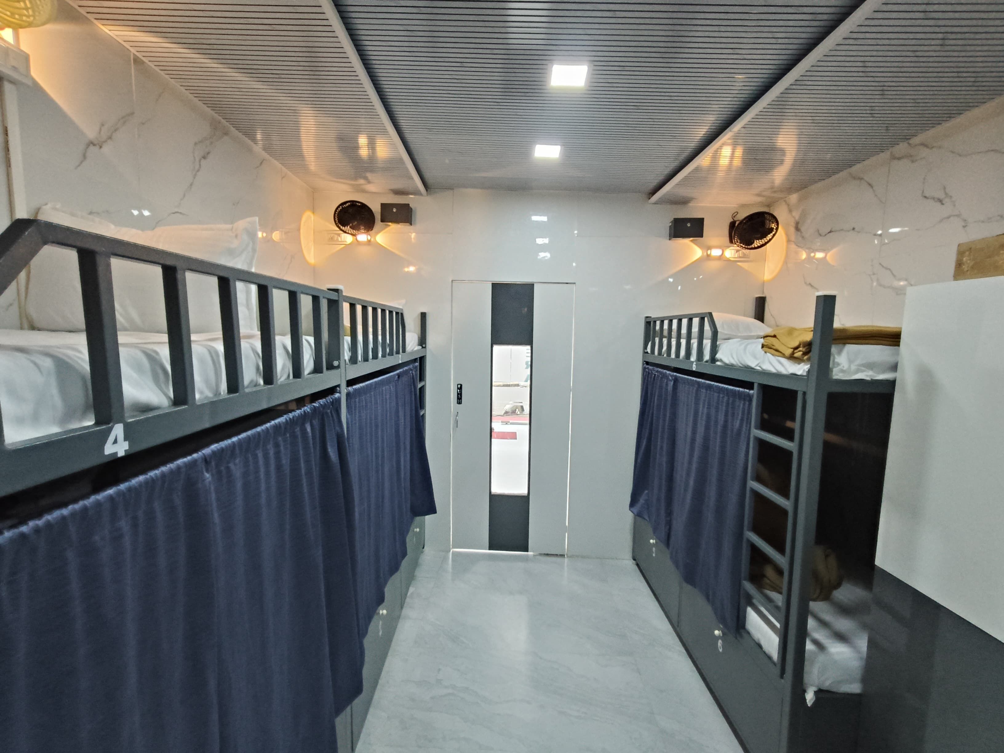 A narrow corridor with bunk beds on either side, a door at the end, and overhead lighting.