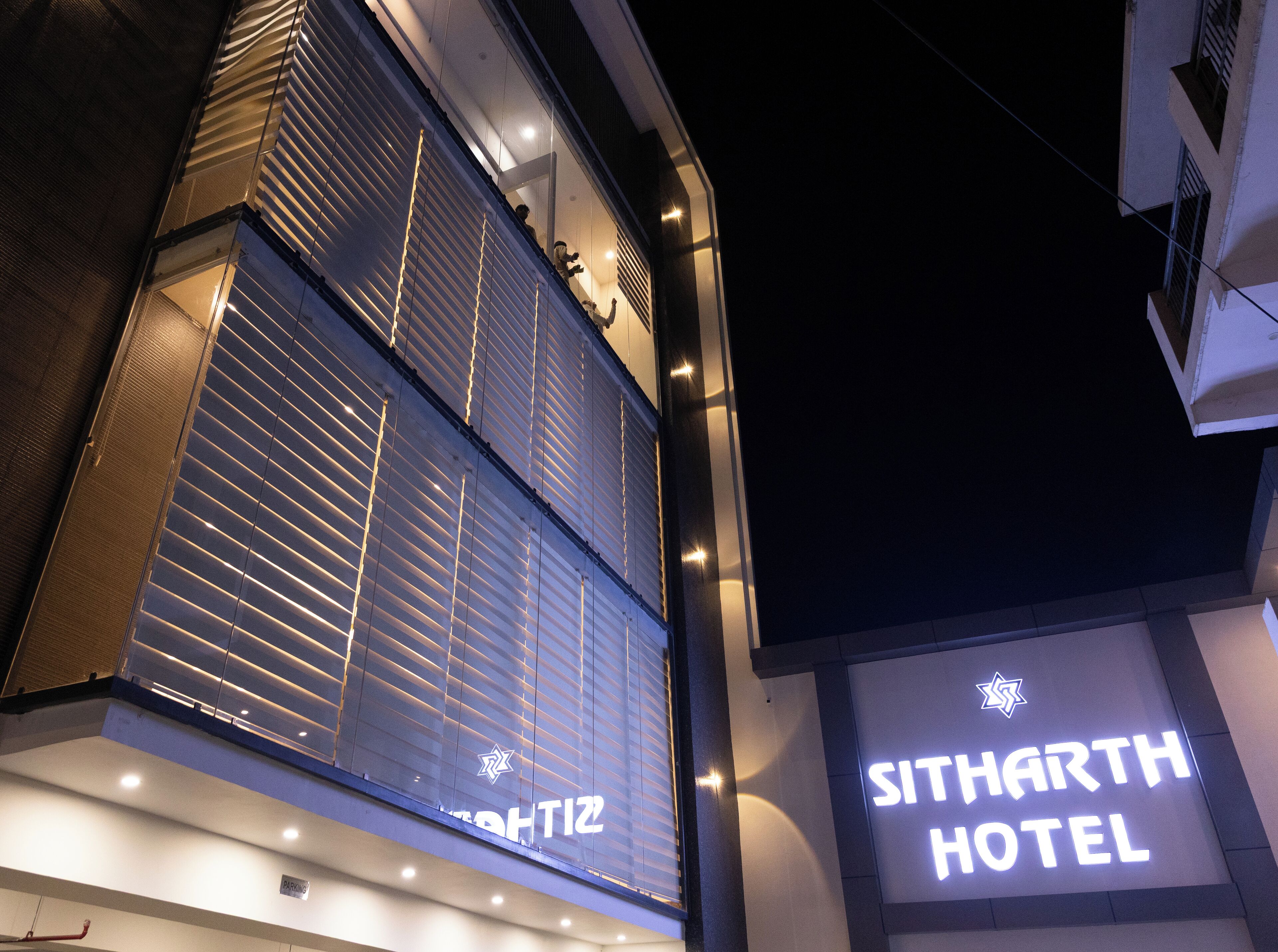 Het uiterlijk van HOTEL SITHARTH, met een opvallend verlicht bord, grote ramen met jaloezieën en een modern architectonisch ontwerp.