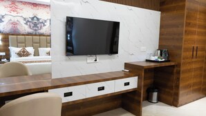 A hotel room with a large flat-screen TV mounted on a marble wall, a wooden desk with drawers, a bed with a headboard, and a trash can. - Sitharth Hotel (Vikasnagar)
