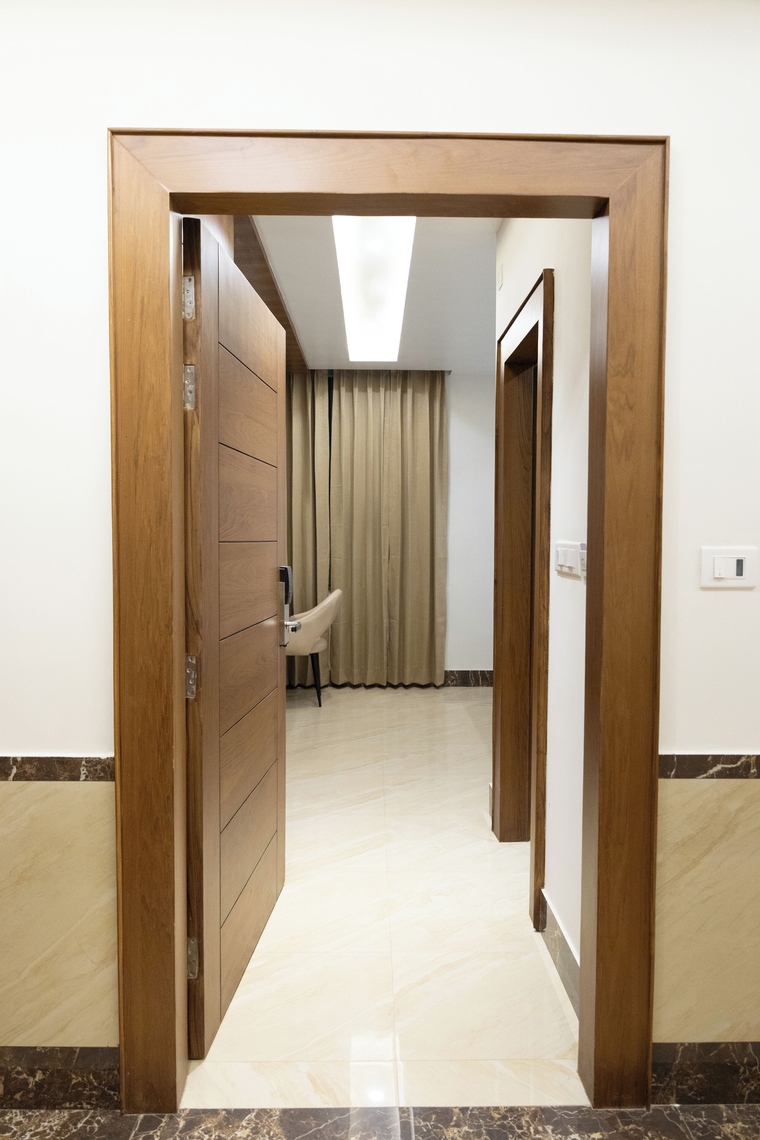 Un couloir avec un cadre de porte en bois, un interrupteur et une chaise au fond.