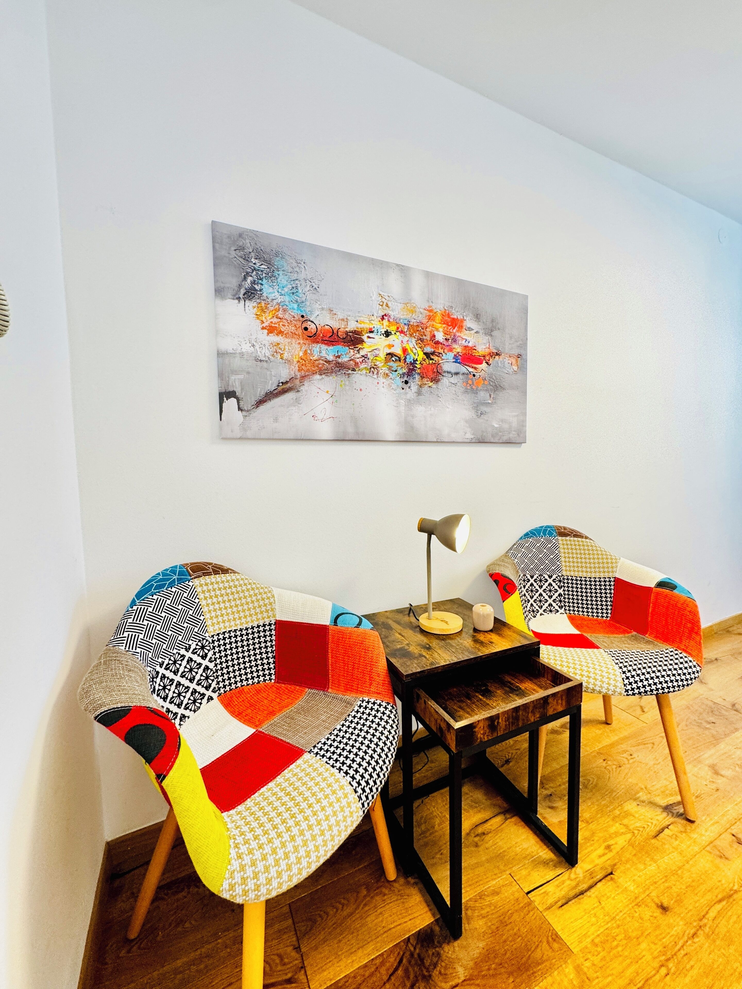 A dining area with two chairs and a table, a painting on the wall, and a lamp on the table.