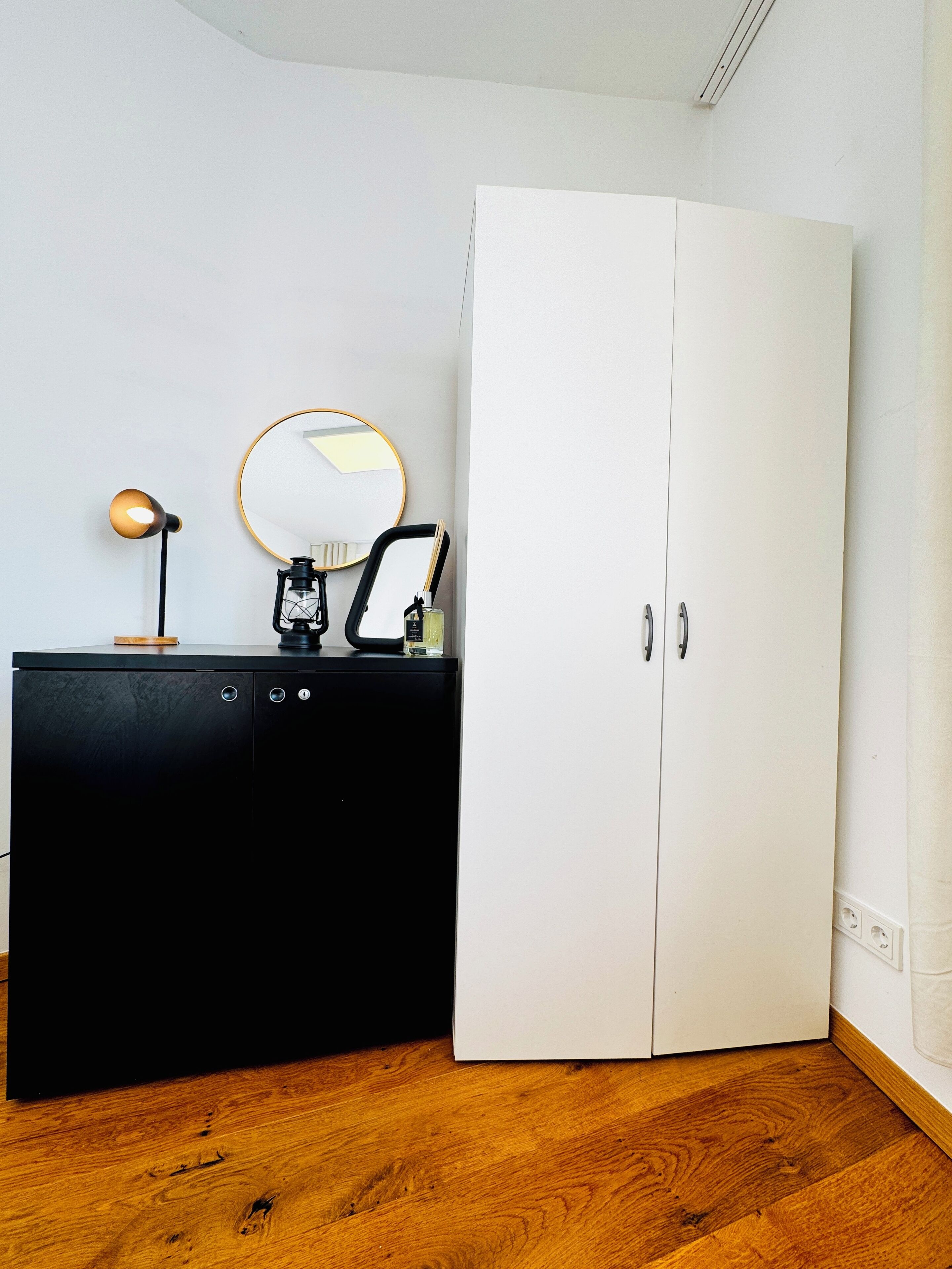 Una habitación con un aparador negro, un armario blanco, un espejo redondo y una lámpara pequeña.
