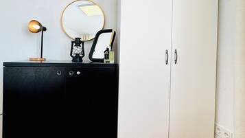 Um quarto com cômoda preta, guarda-roupa branco, espelho redondo e uma pequena luminária.
