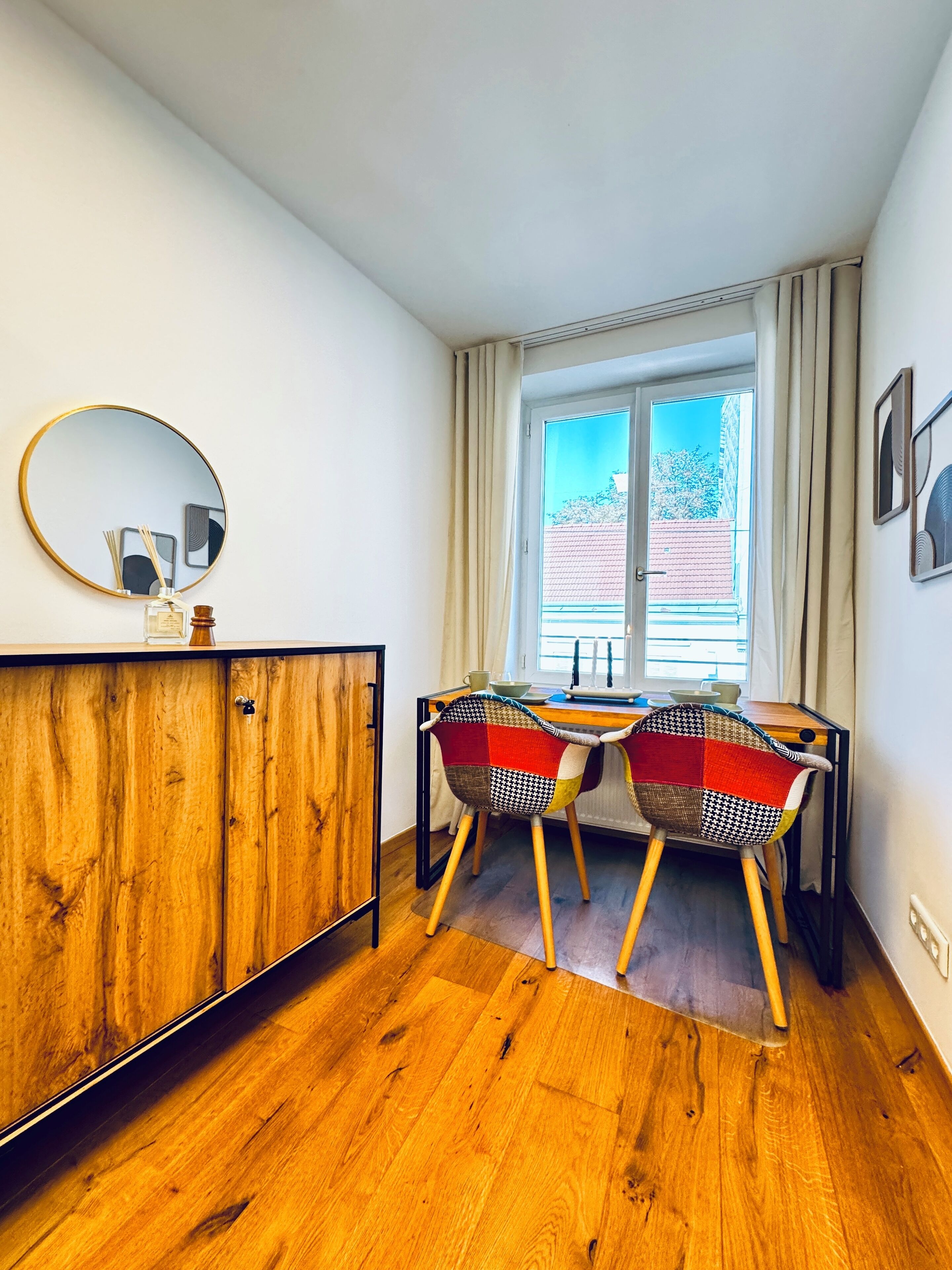 Una habitación con un armario de madera, dos sillas con estampado de cuadros, un espejo redondo y una ventana con cortinas.