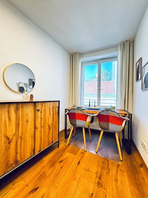 A room with a wooden cabinet, two chairs with a checkered pattern, a round mirror, and a window with curtains.