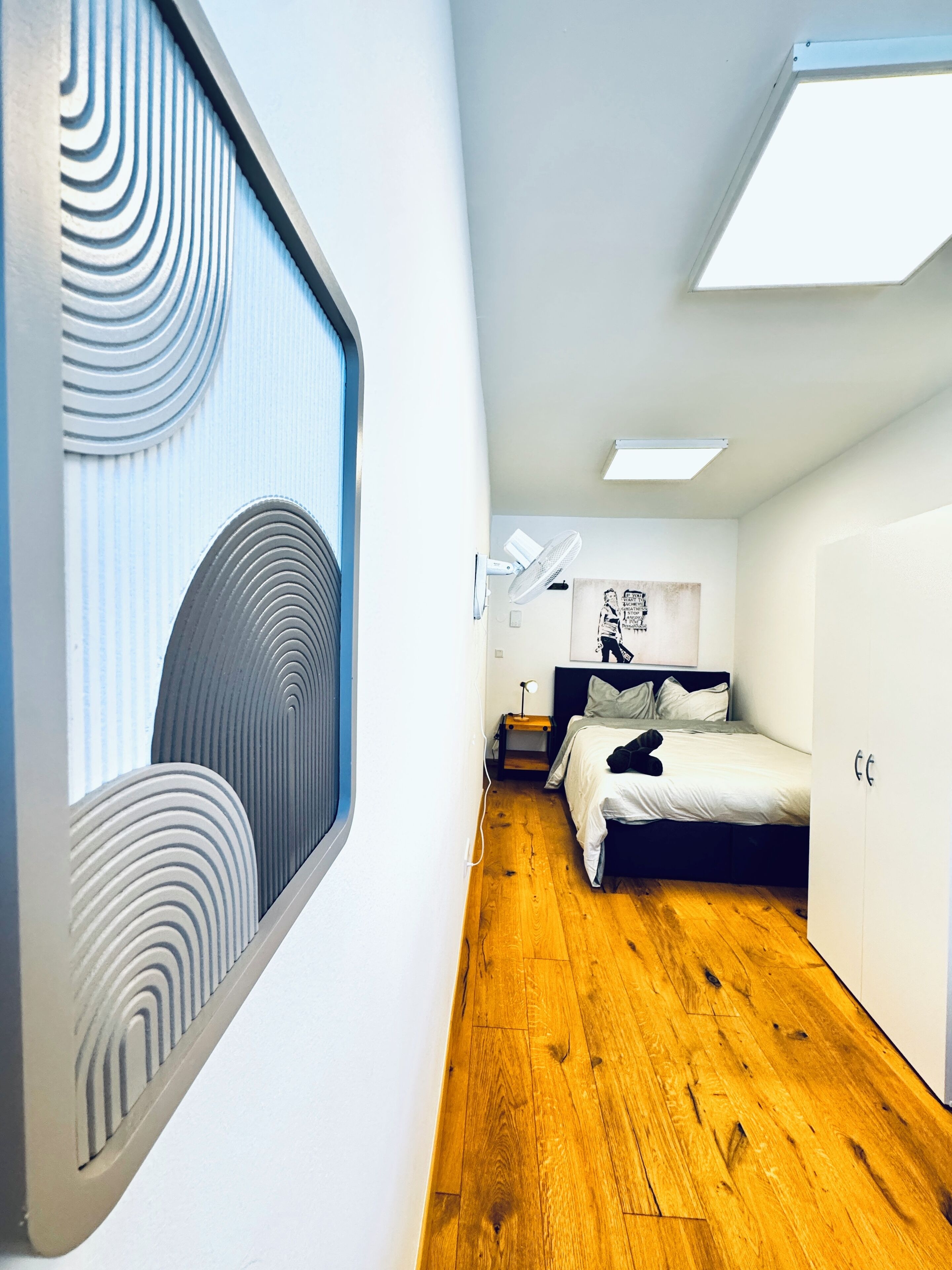 Een moderne slaapkamer met houten vloer, een bed met hoofdbord, een nachtkastje met lamp en een raam met een uniek ontwerp.