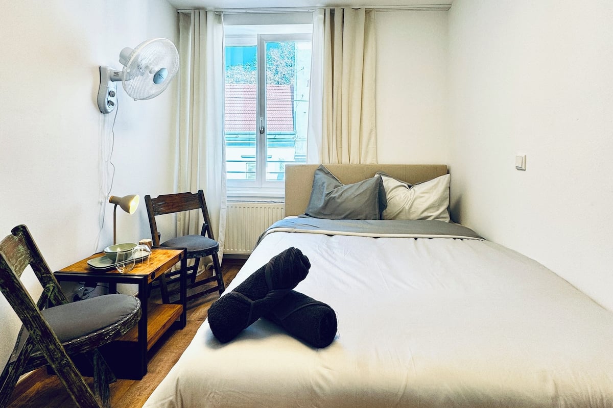 A bedroom with a bed, a desk with a chair, a standing fan, and a window with curtains.