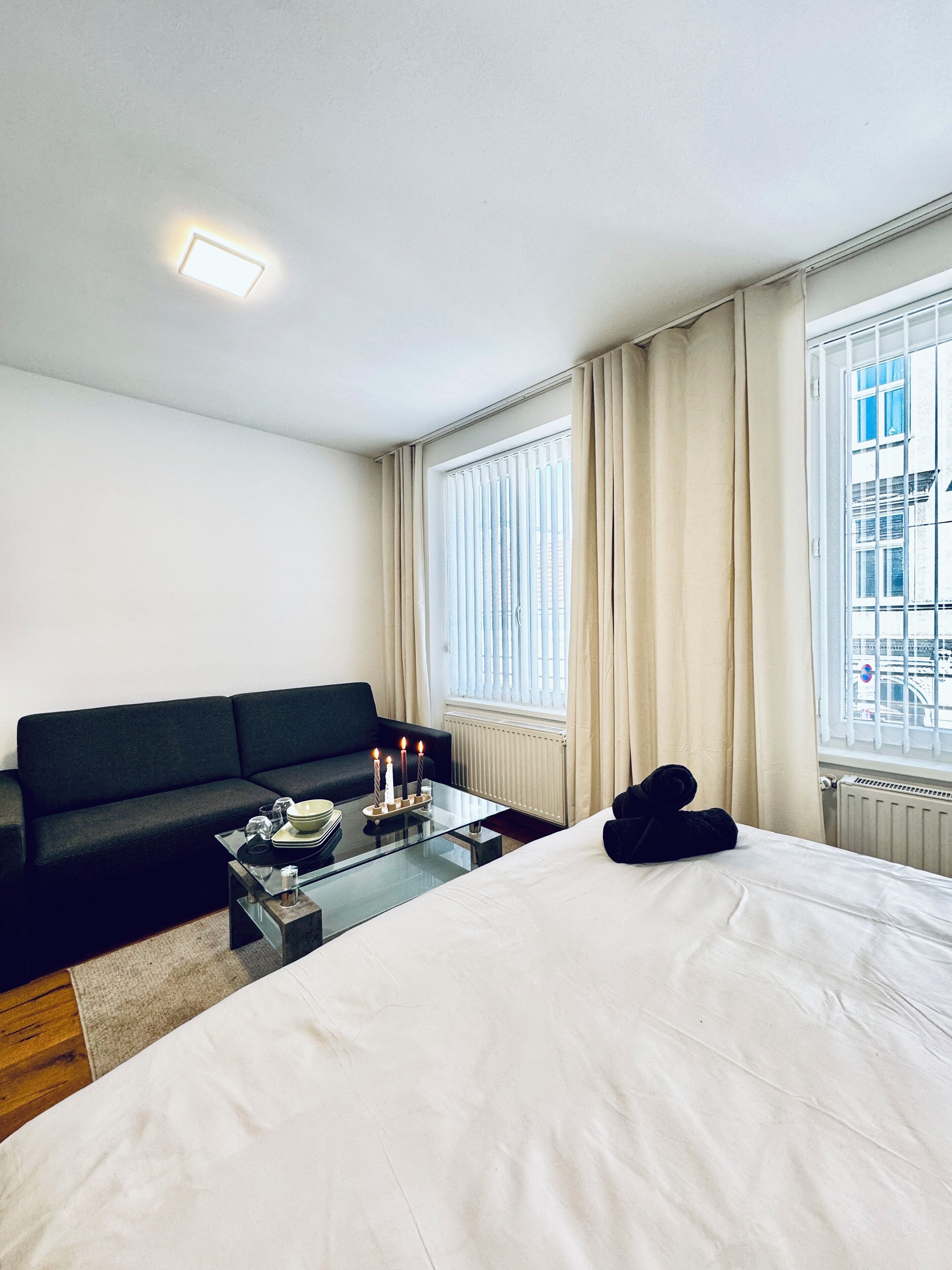 Hotelkamer met een bed, een zwarte bank, een glazen salontafel met kaarsen, en een stadszicht door grote ramen.